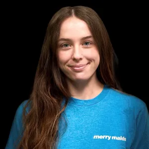 kayla has long brown hair and wearing a blue merry maids tshirt standing in front of a black photo backdrop